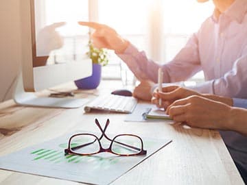 Two people working together at a desk with a computer and glasses
