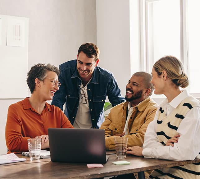 Group of colleagues talking and laughing together around laptop at work