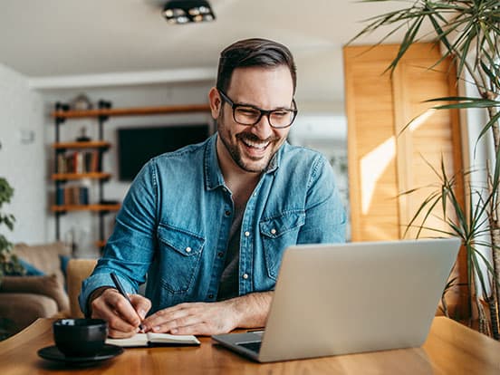 Smiling man in glasses works on a laptop at a wooden table, taking notes in a notebook