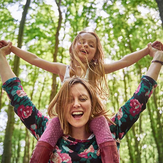 Woman with floral top carries smiling child on shoulders in a sunlit forest. Both have arms outstretched.