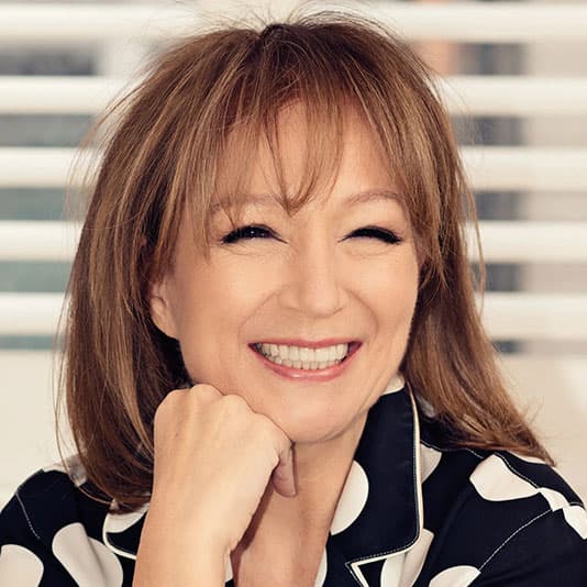 Smiling woman with shoulder-length hair and bangs, wearing a polka dot top, rests her chin on her hand. Blurred striped background.