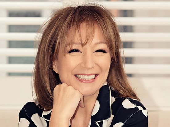 A person with shoulder-length hair smiles warmly, resting their chin on their hand, in front of a background with horizontal blinds.