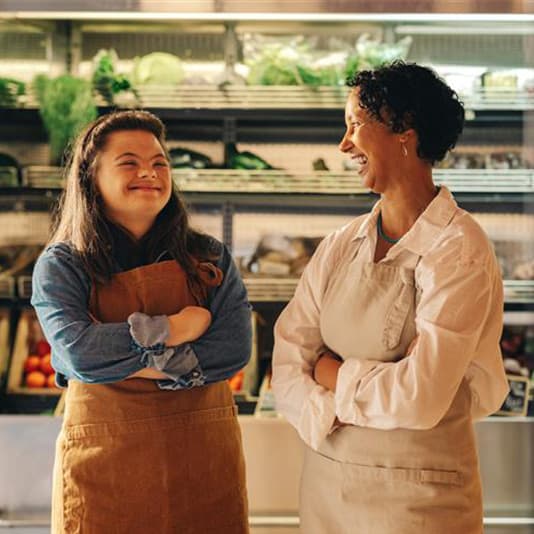 Two people wearing aprons smile at each other in a grocery store, with fresh produce