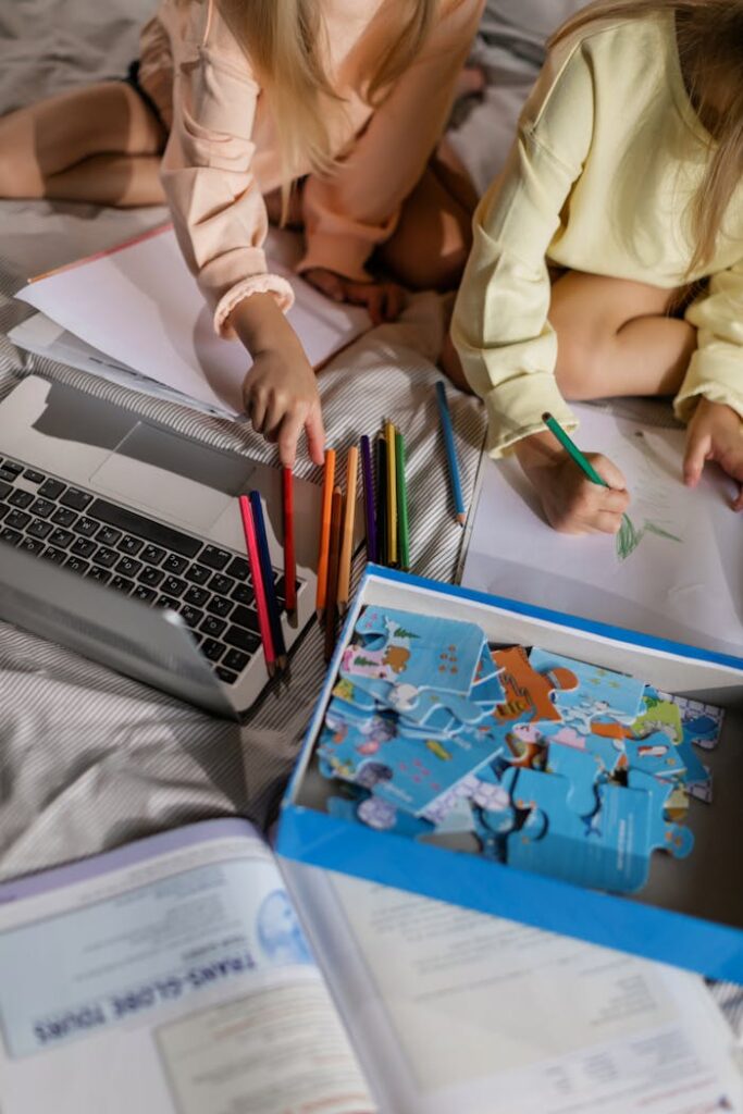 Two children drawing and solving puzzles at home, surrounded by colored pencils and a laptop.